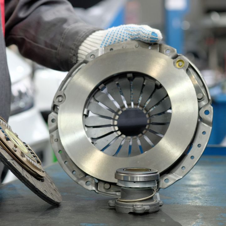 Mechanic holding a clutch pressure plate with a release bearing and clutch disc on a workbench at M & B Parts shop.